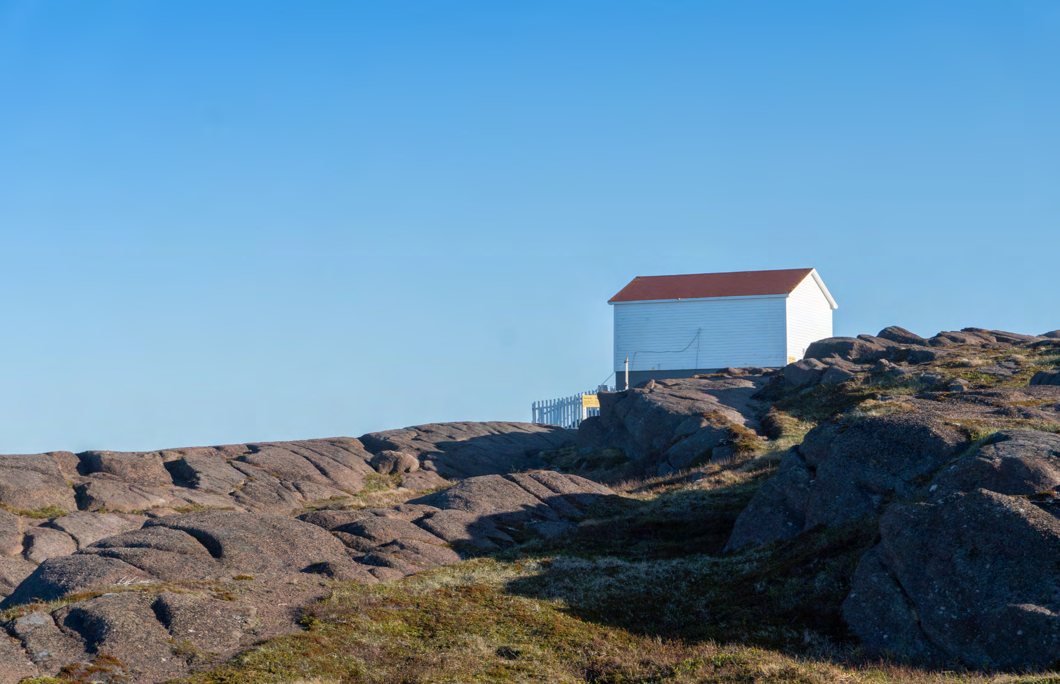 A small white boathouse on a rocky coastal headland under a clear blue sky.